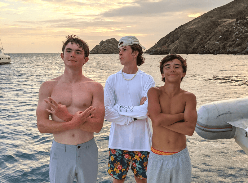 3 teens pose on catamaran with Caribbean island in background on summer dive and sail trip