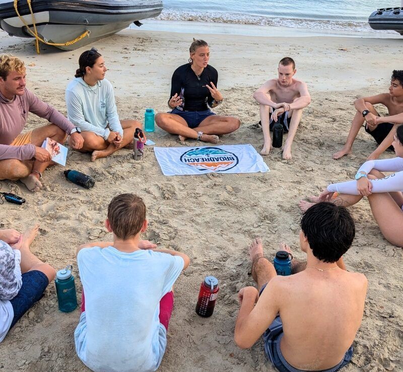 Students sit in circle on beach around Broadreach flag on dive and sail summer program