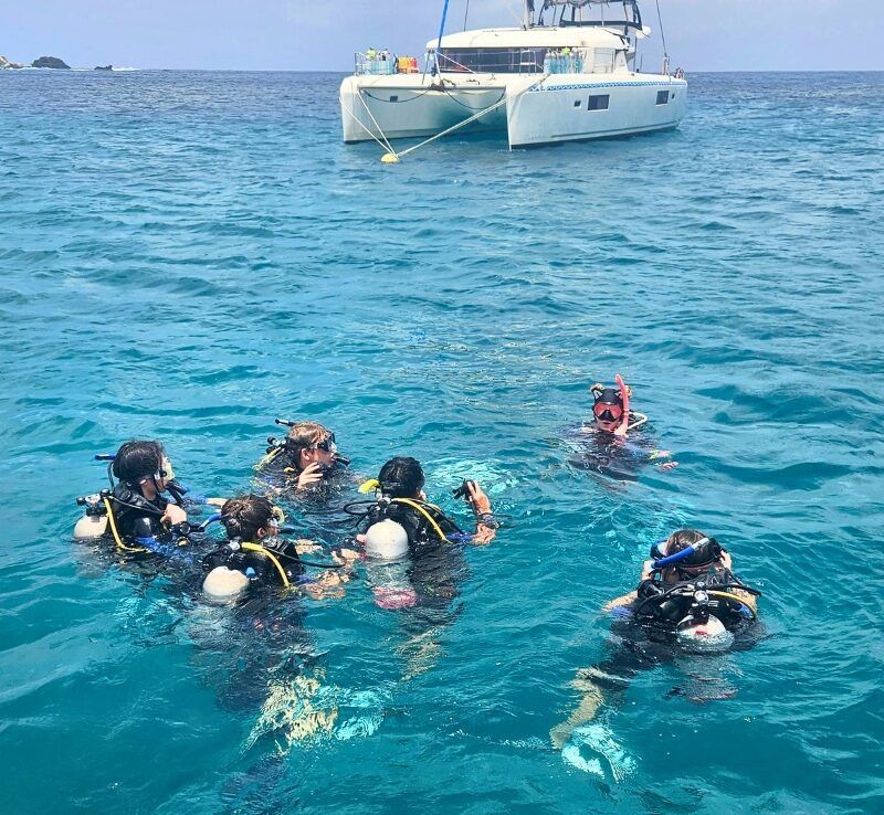 Students in full scuba gear float in the water, listening attentively to a dive instructor giving instructions before a training dive.