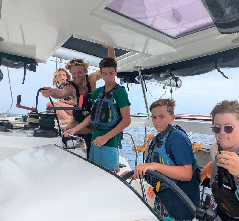 Students work together to sail a catamaran, adjusting lines and steering while an instructor offers direction nearby.