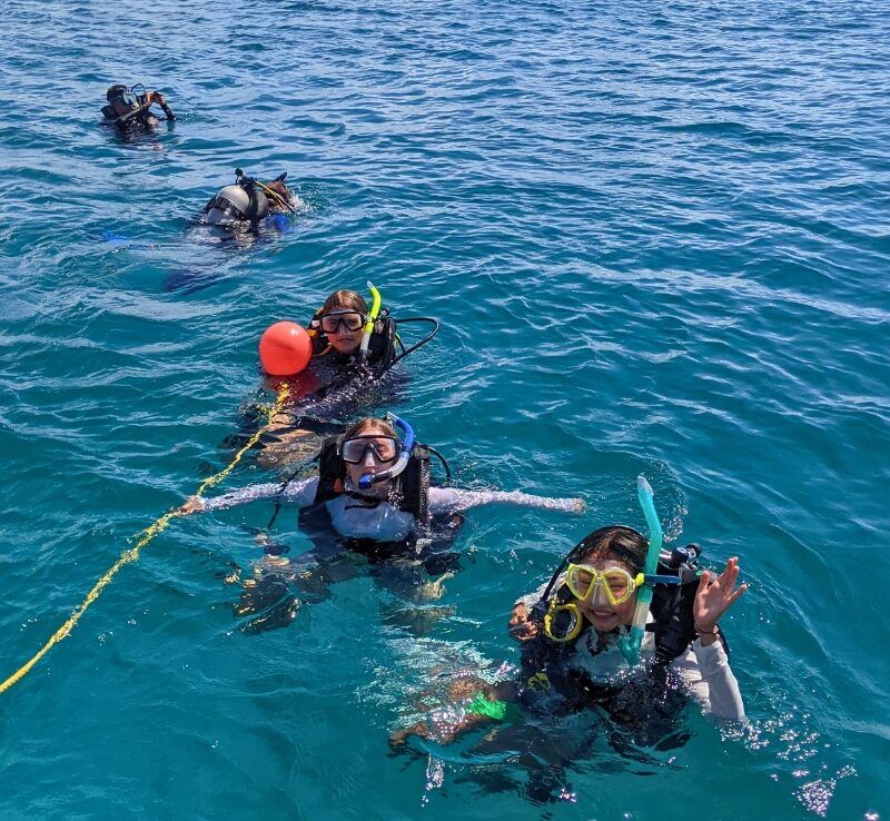 A group of middle school students in scuba gear float on the surface, holding onto a line as they get ready to begin their dive.