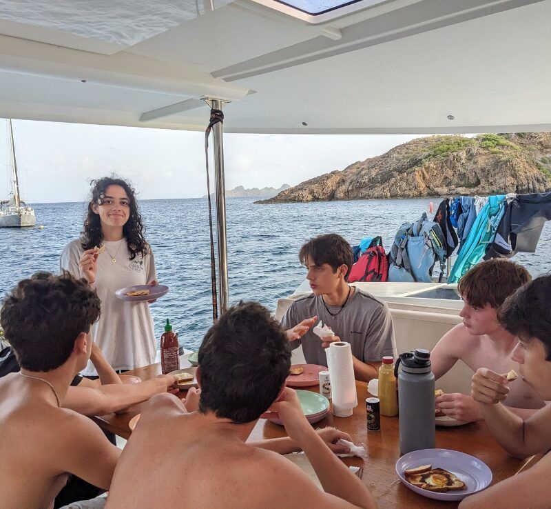 A group of high school students sit around a table on a catamaran, enjoying a meal together with the ocean in the background.