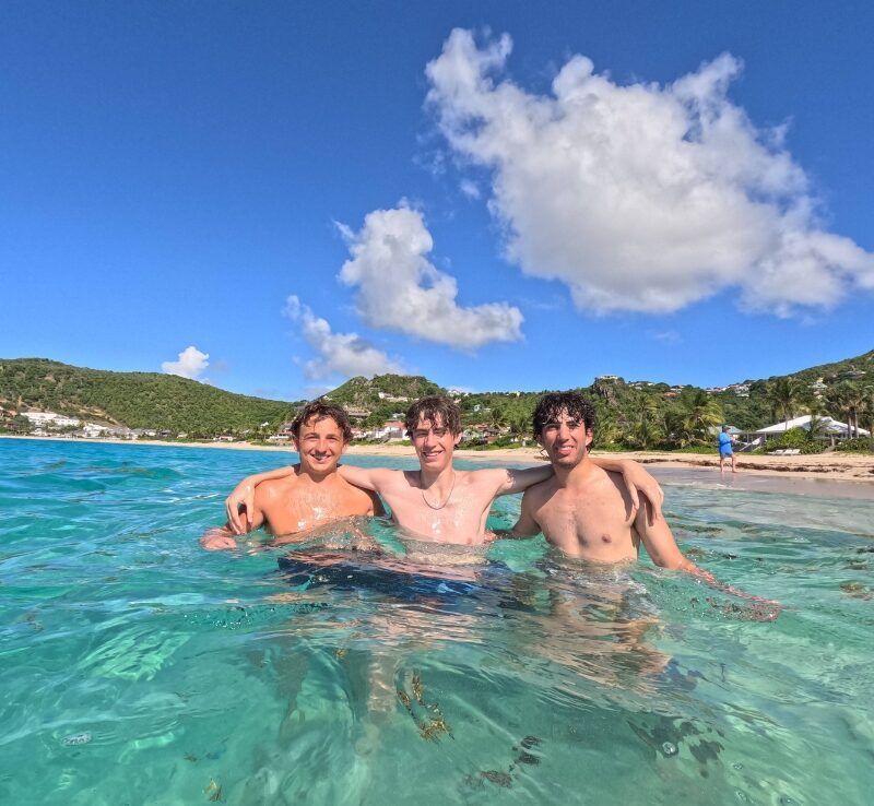 Three high school boys stand in shallow, clear water near a sandy Caribbean beach on a sunny day.