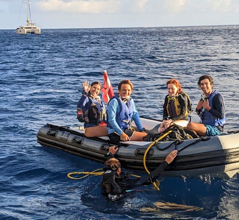 A group of high school students smile and laugh while riding together on a dinghy in calm Caribbean waters.