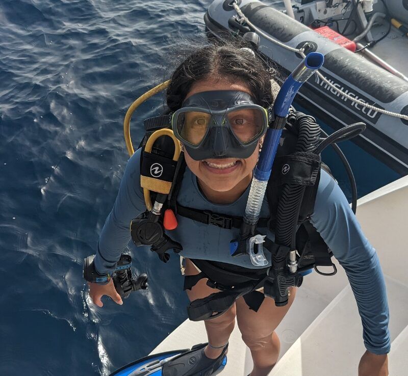 A girl in scuba gear stands on the edge of a catamaran, looking up at the camera with a smile before a dive.