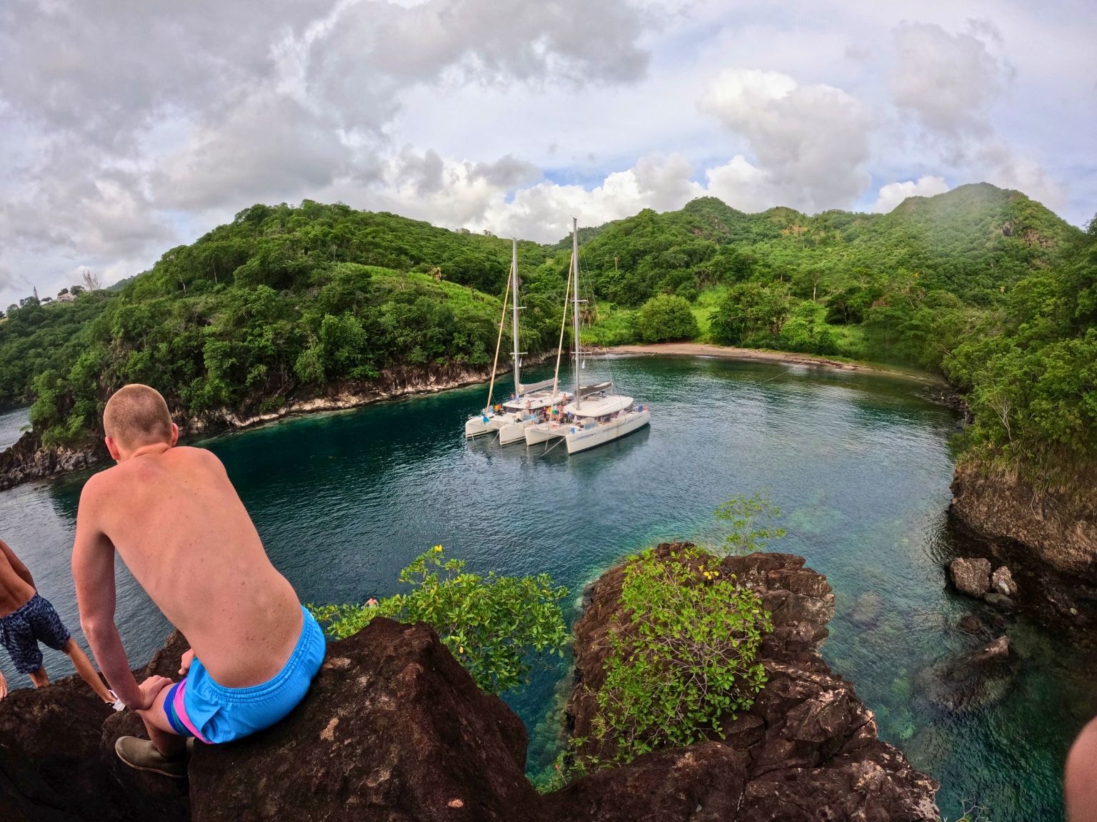 Teen sitting on a rocky cliff overlooking a catamaran anchored in a lush, tropical cove in St. Vincent